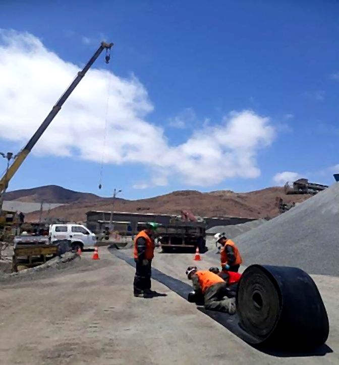 Personal en terreno extendiendo un rollo de correa transportadora con apoyo de grúa